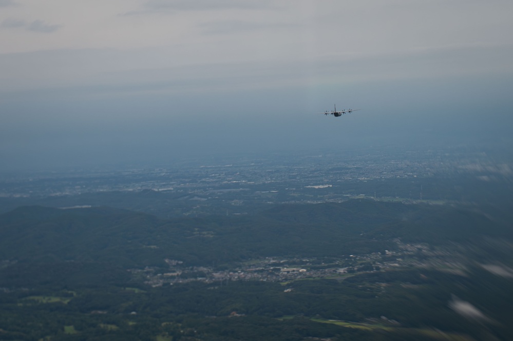 An aerial photograph taken from altitude showing a military transport aircraft, consistent with a C-130 Hercules silhouette, flying over a vast landscape of mixed terrain including forested hills, urban settlements, and open plains. The image is captured from another airborne platform looking outward at the subject aircraft. The overall scene suggests a military flight operation over what appears to be a densely populated region.