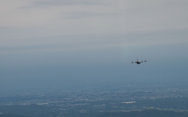 An aerial photograph taken from altitude showing a military transport aircraft, consistent with a C-130 Hercules silhouette, flying over a vast landscape of mixed terrain including forested hills, urban settlements, and open plains. The image is captured from another airborne platform looking outward at the subject aircraft. The overall scene suggests a military flight operation over what appears to be a densely populated region.