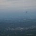 An aerial photograph taken from altitude showing a military transport aircraft, consistent with a C-130 Hercules silhouette, flying over a vast landscape of mixed terrain including forested hills, urban settlements, and open plains. The image is captured from another airborne platform looking outward at the subject aircraft. The overall scene suggests a military flight operation over what appears to be a densely populated region.