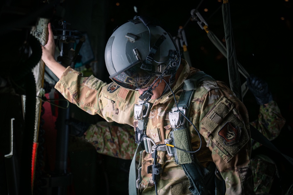 A military personnel member wearing full tactical gear, including a flight helmet with visor, multicam/OCP camouflage uniform, and a parachute harness system, is positioned inside what appears to be a military aircraft. The individual is reaching upward with one hand, likely preparing for or conducting a parachute jump operation. Patches on the uniform suggest affiliation with the United States Air Force.