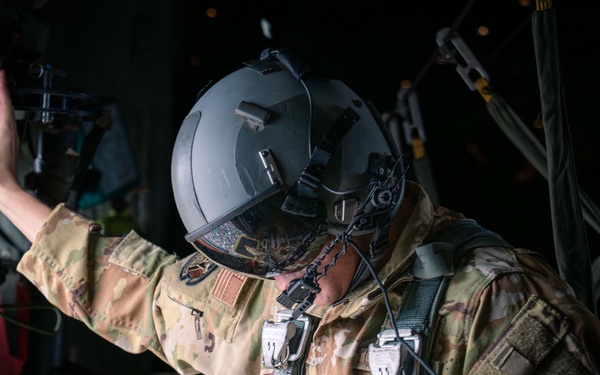A military personnel member wearing full tactical gear, including a flight helmet with visor, multicam/OCP camouflage uniform, and a parachute harness system, is positioned inside what appears to be a military aircraft. The individual is reaching upward with one hand, likely preparing for or conducting a parachute jump operation. Patches on the uniform suggest affiliation with the United States Air Force.