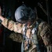 A military personnel member wearing full tactical gear, including a flight helmet with visor, multicam/OCP camouflage uniform, and a parachute harness system, is positioned inside what appears to be a military aircraft. The individual is reaching upward with one hand, likely preparing for or conducting a parachute jump operation. Patches on the uniform suggest affiliation with the United States Air Force.