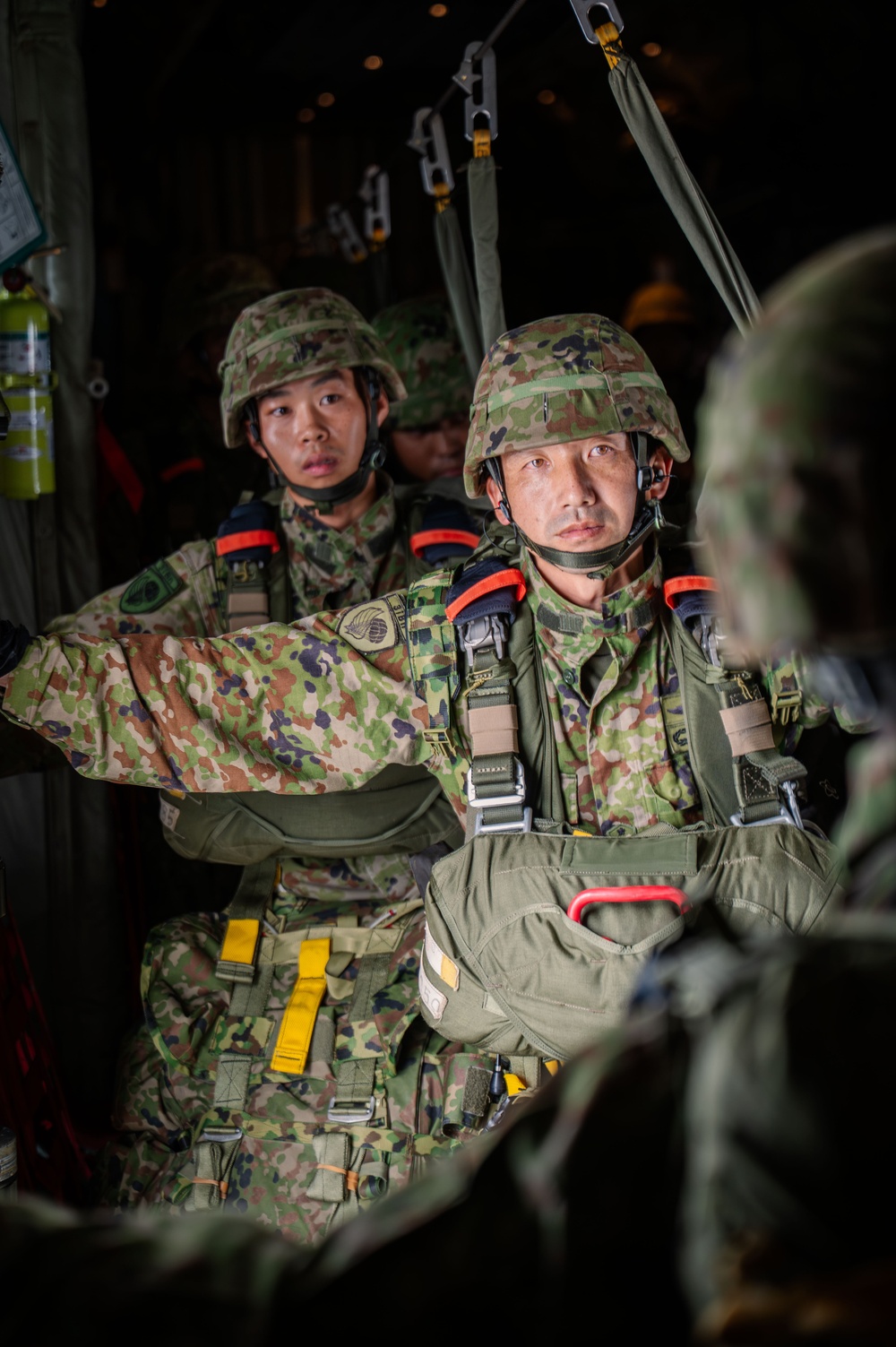Two soldiers wearing Japanese Ground Self-Defense Force (JGSDF) camouflage uniforms and helmets are seated inside a military aircraft, equipped with parachute harnesses and jump gear. They appear to be preparing for an airborne operation, with static lines visible overhead. The foreground soldier has a focused, composed expression while the background soldier appears alert.