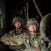 Two soldiers wearing Japanese Ground Self-Defense Force (JGSDF) camouflage uniforms and helmets are seated inside a military aircraft, equipped with parachute harnesses and jump gear. They appear to be preparing for an airborne operation, with static lines visible overhead. The foreground soldier has a focused, composed expression while the background soldier appears alert.