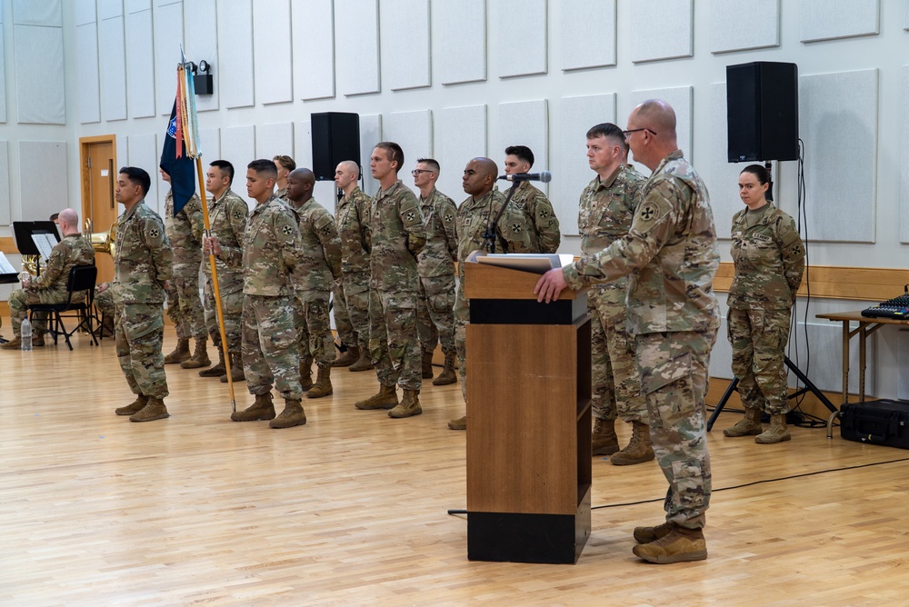 A military ceremony is taking place in an indoor hall with hardwood floors and acoustic wall panels. A senior soldier stands at a podium addressing a formation of approximately 15 soldiers in OCP (Operational Camouflage Pattern) uniforms, with one soldier carrying a unit guidon. A military band is partially visible on the left side of the image.