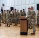 A military ceremony is taking place in an indoor hall with hardwood floors and acoustic wall panels. A senior soldier stands at a podium addressing a formation of approximately 15 soldiers in OCP (Operational Camouflage Pattern) uniforms, with one soldier carrying a unit guidon. A military band is partially visible on the left side of the image.