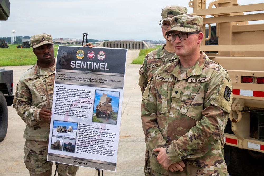 Three U.S. Army soldiers are gathered outdoors near military vehicles for a briefing or demonstration involving the 'SENTINEL' air defense system. A presentation board labeled 'SENTINEL' and marked 'UNCLASSIFIED' is displayed on an easel, describing advanced air defense capabilities. The soldiers are in OCP (Operational Camouflage Pattern) uniforms with visible name tapes and unit patches.