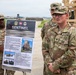 Three U.S. Army soldiers are gathered outdoors near military vehicles for a briefing or demonstration involving the 'SENTINEL' air defense system. A presentation board labeled 'SENTINEL' and marked 'UNCLASSIFIED' is displayed on an easel, describing advanced air defense capabilities. The soldiers are in OCP (Operational Camouflage Pattern) uniforms with visible name tapes and unit patches.