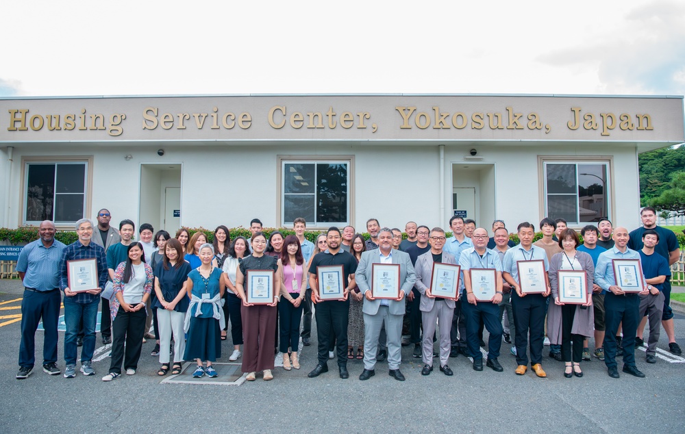 A large group of approximately 35-40 people are posed for a formal group photograph in front of the Housing Service Center in Yokosuka, Japan. Several individuals in the front rows are holding framed certificates or plaques, suggesting an awards ceremony or recognition event. The setting appears to be a U.S. military installation based on the facility name and context.
