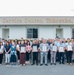 A large group of approximately 35-40 people are posed for a formal group photograph in front of the Housing Service Center in Yokosuka, Japan. Several individuals in the front rows are holding framed certificates or plaques, suggesting an awards ceremony or recognition event. The setting appears to be a U.S. military installation based on the facility name and context.