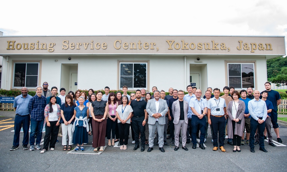 A large group of approximately 40-50 people pose for a formal group photograph in front of the Housing Service Center in Yokosuka, Japan. The group appears to be a mix of staff members and officials of diverse backgrounds, dressed in business and business-casual attire. The building's signage is clearly visible above the group.