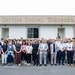 A large group of approximately 40-50 people pose for a formal group photograph in front of the Housing Service Center in Yokosuka, Japan. The group appears to be a mix of staff members and officials of diverse backgrounds, dressed in business and business-casual attire. The building's signage is clearly visible above the group.