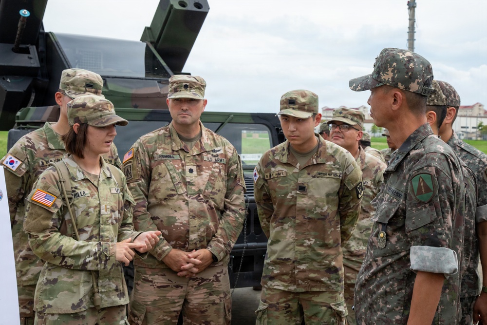A group of U.S. Army soldiers and what appears to be a Republic of Korea (ROK) Army officer are engaged in a briefing or discussion outdoors near military vehicles. A female U.S. Army soldier appears to be presenting or explaining something, while others listen attentively. The setting suggests a joint military exercise or equipment demonstration.