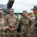 A group of U.S. Army soldiers and what appears to be a Republic of Korea (ROK) Army officer are engaged in a briefing or discussion outdoors near military vehicles. A female U.S. Army soldier appears to be presenting or explaining something, while others listen attentively. The setting suggests a joint military exercise or equipment demonstration.