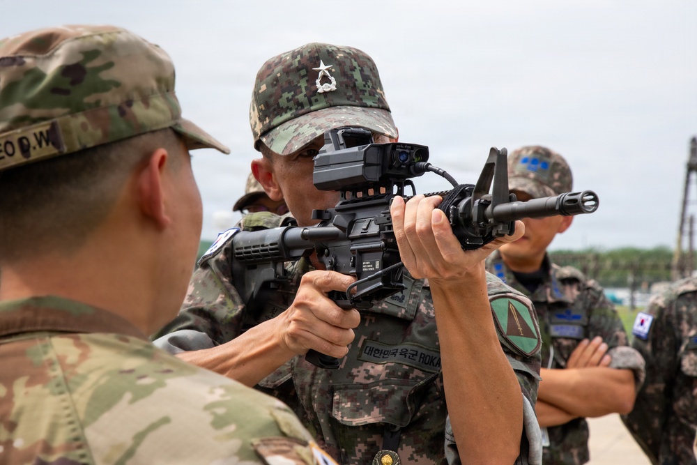 The image shows military personnel from the Republic of Korea Army (ROKA) and what appears to be the U.S. Army engaged in a weapons familiarization or training exercise. A ROKA soldier is aiming an M4-style carbine equipped with an advanced optic/sight system while a U.S. soldier observes. Additional ROKA soldiers are visible in the background, suggesting a joint military training event.