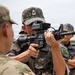 The image shows military personnel from the Republic of Korea Army (ROKA) and what appears to be the U.S. Army engaged in a weapons familiarization or training exercise. A ROKA soldier is aiming an M4-style carbine equipped with an advanced optic/sight system while a U.S. soldier observes. Additional ROKA soldiers are visible in the background, suggesting a joint military training event.