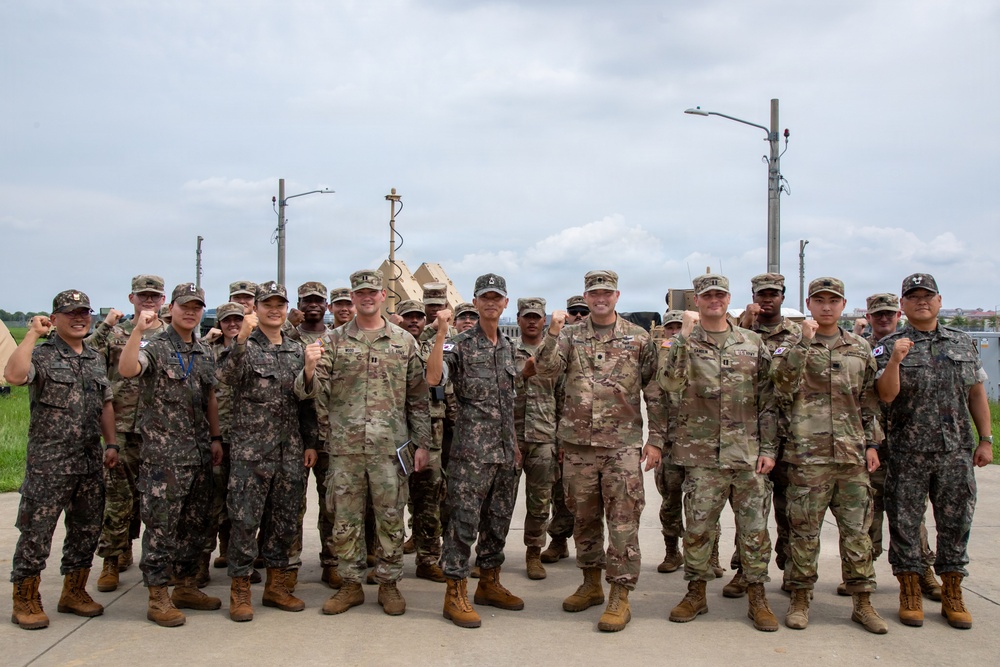 A group of approximately 20-25 military personnel from two distinct forces are posing together for a group photo outdoors. The soldiers are wearing two different camouflage uniform patterns, consistent with U.S. Army MultiCam/OCP and Republic of Korea (ROK) Army digital camouflage uniforms. Many individuals are raising their fists in a unified gesture, suggesting a joint military exercise or cooperative event.