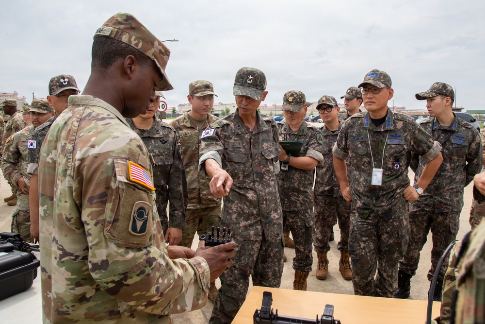A U.S. Army soldier is conducting a demonstration or briefing for a group of Republic of Korea (ROK) Army personnel at what appears to be a joint military exercise or equipment showcase. The U.S. soldier, wearing OCP (Operational Camouflage Pattern) uniform with an American flag patch, is presenting a piece of equipment to attentive ROK military officers and soldiers. This appears to be a bilateral military cooperation event between U.S. and South Korean forces.