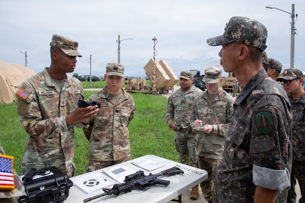 A group of U.S. Army soldiers and what appear to be Republic of Korea (ROK) Army personnel are engaged in a weapons familiarization or demonstration briefing outdoors. A soldier identified by nametape as 'Williams' is holding a handgun and appears to be explaining or demonstrating it to the group. Military equipment, including what appears to be a PATRIOT missile defense system, is visible in the background.