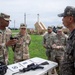 A group of U.S. Army soldiers and what appear to be Republic of Korea (ROK) Army personnel are engaged in a weapons familiarization or demonstration briefing outdoors. A soldier identified by nametape as 'Williams' is holding a handgun and appears to be explaining or demonstrating it to the group. Military equipment, including what appears to be a PATRIOT missile defense system, is visible in the background.