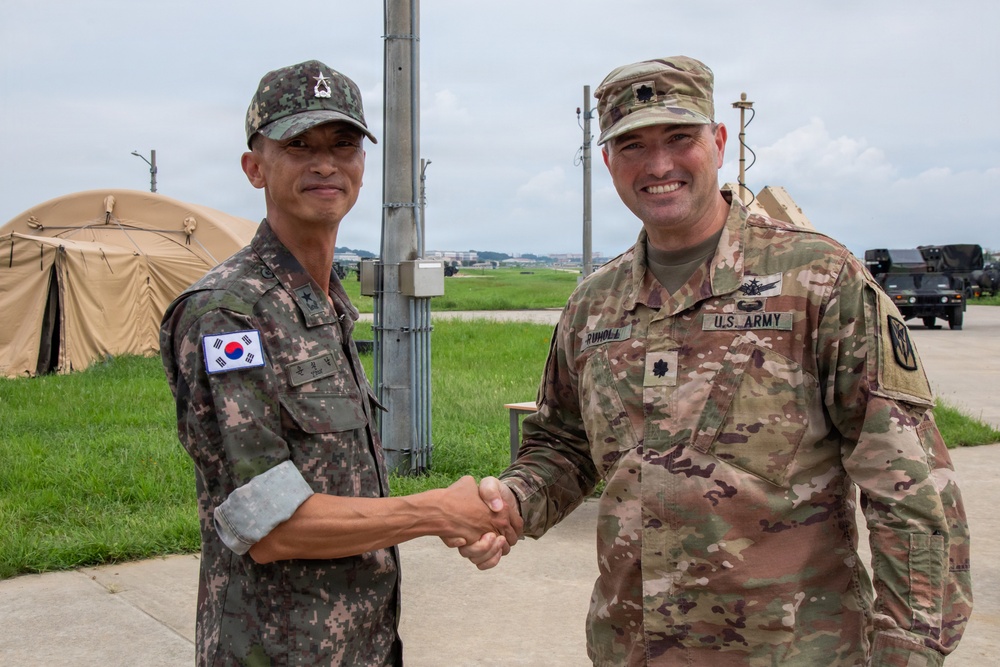 Two military officers, one from the Republic of Korea (ROK) Army and one from the U.S. Army, are shaking hands in what appears to be a joint military exercise or base setting. The U.S. soldier wears the rank of Lieutenant Colonel (oak leaf insignia) and has a name tape reading 'RUHOLL' with 'U.S. ARMY' visible on his uniform. The setting includes military tents and vehicles in the background, suggesting a field operations environment.