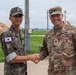 Two military officers, one from the Republic of Korea (ROK) Army and one from the U.S. Army, are shaking hands in what appears to be a joint military exercise or base setting. The U.S. soldier wears the rank of Lieutenant Colonel (oak leaf insignia) and has a name tape reading 'RUHOLL' with 'U.S. ARMY' visible on his uniform. The setting includes military tents and vehicles in the background, suggesting a field operations environment.
