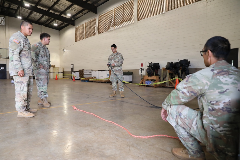 Four U.S. Army soldiers in MultiCam/OCP camouflage uniforms are gathered inside a large indoor facility, likely a gymnasium or warehouse, engaged in what appears to be a rope or rigging training exercise. One soldier in the center is holding a dark rope/cord while a red rope or strap is laid out on the concrete floor. Two soldiers observe from the left while a fourth crouches on the right.