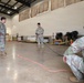 Four U.S. Army soldiers in MultiCam/OCP camouflage uniforms are gathered inside a large indoor facility, likely a gymnasium or warehouse, engaged in what appears to be a rope or rigging training exercise. One soldier in the center is holding a dark rope/cord while a red rope or strap is laid out on the concrete floor. Two soldiers observe from the left while a fourth crouches on the right.