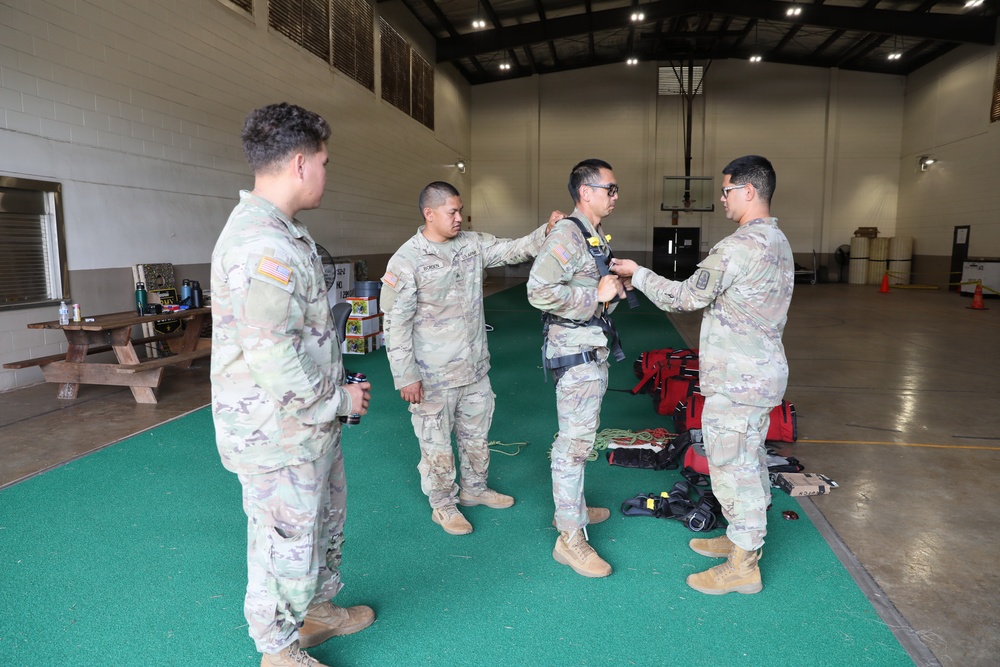 Four U.S. Army soldiers in OCP (Operational Camouflage Pattern) uniforms are gathered inside what appears to be a military gymnasium or large indoor facility. One soldier is being fitted or inspected with a harness/rappelling equipment by another soldier, while two others observe. Various pieces of rescue or rappelling gear are visible on the floor nearby.