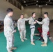 Four U.S. Army soldiers in OCP (Operational Camouflage Pattern) uniforms are gathered inside what appears to be a military gymnasium or large indoor facility. One soldier is being fitted or inspected with a harness/rappelling equipment by another soldier, while two others observe. Various pieces of rescue or rappelling gear are visible on the floor nearby.