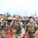 A group of U.S. Army soldiers in MultiCam (OCP) uniforms and plate carriers are conducting a litter carry drill on a red running track. They are transporting a training mannequin on a military stretcher/litter as part of what appears to be a combat casualty care or physical fitness exercise. Multiple soldiers wearing 2nd Infantry Division patches are visible, with additional personnel participating in the background.