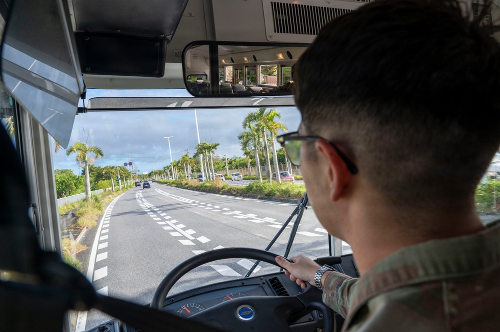 A military service member in camouflage uniform is driving a large bus along a palm tree-lined road, likely in a tropical location such as Okinawa, Japan. The view is from behind the driver, showing the windshield perspective of a multi-lane road with light traffic. The interior includes a large rearview mirror mounted above the windshield and a steering wheel with a blue emblem.