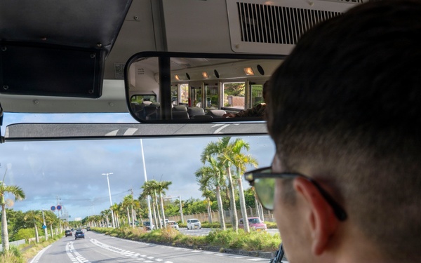 A military service member in camouflage uniform is driving a large bus along a palm tree-lined road, likely in a tropical location such as Okinawa, Japan. The view is from behind the driver, showing the windshield perspective of a multi-lane road with light traffic. The interior includes a large rearview mirror mounted above the windshield and a steering wheel with a blue emblem.