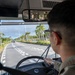 A military service member in camouflage uniform is driving a large bus along a palm tree-lined road, likely in a tropical location such as Okinawa, Japan. The view is from behind the driver, showing the windshield perspective of a multi-lane road with light traffic. The interior includes a large rearview mirror mounted above the windshield and a steering wheel with a blue emblem.