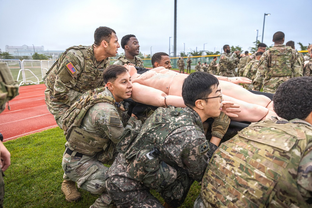 Military personnel in mixed uniforms, including US Army OCP camouflage and what appears to be Republic of Korea Army digital camouflage, are conducting a joint training exercise. They are collectively carrying a life-sized medical training mannequin on a litter/stretcher across a grass field adjacent to a running track. This appears to be a combined arms or allied forces casualty evacuation (CASEVAC) training drill.