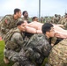 Military personnel in mixed uniforms, including US Army OCP camouflage and what appears to be Republic of Korea Army digital camouflage, are conducting a joint training exercise. They are collectively carrying a life-sized medical training mannequin on a litter/stretcher across a grass field adjacent to a running track. This appears to be a combined arms or allied forces casualty evacuation (CASEVAC) training drill.