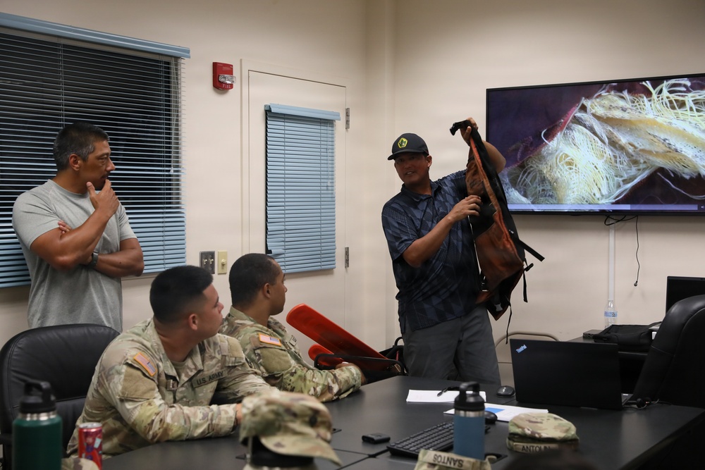 A briefing or training session is taking place in a military classroom setting. A civilian instructor is holding up what appears to be a damaged or worn backpack/gear item while presenting to U.S. Army soldiers seated at a table. A large display screen behind him shows a close-up image of tangled or damaged material, likely related to the presentation topic.