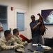 A briefing or training session is taking place in a military classroom setting. A civilian instructor is holding up what appears to be a damaged or worn backpack/gear item while presenting to U.S. Army soldiers seated at a table. A large display screen behind him shows a close-up image of tangled or damaged material, likely related to the presentation topic.