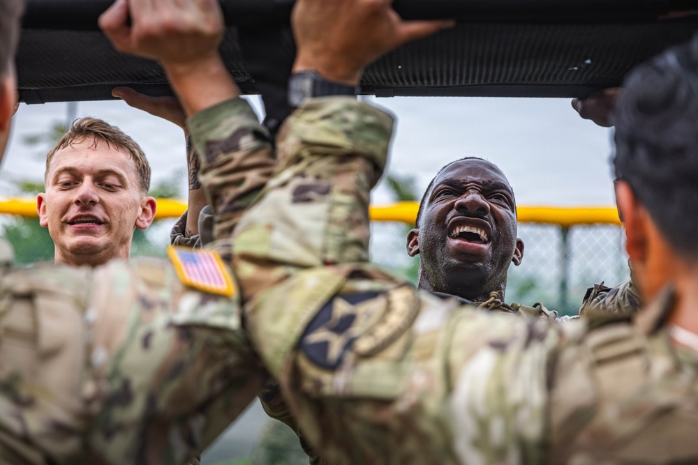U.S. Army soldiers in MultiCam (OCP) uniforms are performing pull-ups or a similar overhead bar exercise during what appears to be a physical fitness training event. The soldiers display intense physical exertion, with visible strain and determination on their faces. The setting appears to be an outdoor military fitness course, likely in a humid or rainy environment based on the wet appearance of the soldiers.