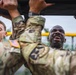 U.S. Army soldiers in MultiCam (OCP) uniforms are performing pull-ups or a similar overhead bar exercise during what appears to be a physical fitness training event. The soldiers display intense physical exertion, with visible strain and determination on their faces. The setting appears to be an outdoor military fitness course, likely in a humid or rainy environment based on the wet appearance of the soldiers.