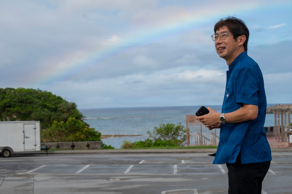 A middle-aged man wearing a blue short-sleeve shirt and glasses stands in a parking lot near a coastal area. He is holding a smartphone and wearing a wristwatch, smiling while looking to his left. A rainbow is visible in the cloudy sky above the ocean in the background.
