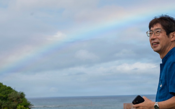 A middle-aged man wearing a blue short-sleeve shirt and glasses stands in a parking lot near a coastal area. He is holding a smartphone and wearing a wristwatch, smiling while looking to his left. A rainbow is visible in the cloudy sky above the ocean in the background.