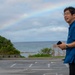 A middle-aged man wearing a blue short-sleeve shirt and glasses stands in a parking lot near a coastal area. He is holding a smartphone and wearing a wristwatch, smiling while looking to his left. A rainbow is visible in the cloudy sky above the ocean in the background.