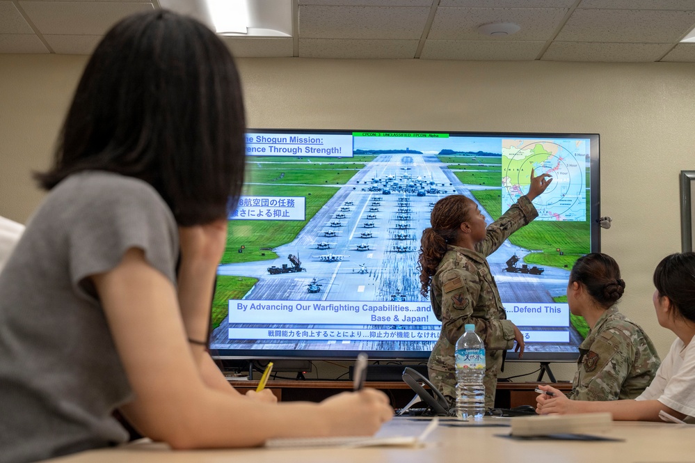 A military briefing is taking place in a conference room where a U.S. Air Force service member in camouflage uniform is presenting a slide to a mixed audience of military personnel and civilians. The presentation screen displays a slide titled 'The Shogun Mission: Deterrence Through Strength' with bilingual English and Japanese text, showing an airfield with military aircraft and a regional map. The presenter is pointing to a map in the upper right corner of the screen that appears to show East Asia with range circles.