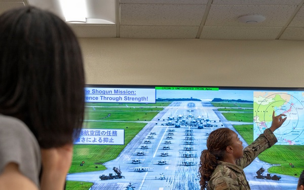 A military briefing is taking place in a conference room where a U.S. Air Force service member in camouflage uniform is presenting a slide to a mixed audience of military personnel and civilians. The presentation screen displays a slide titled 'The Shogun Mission: Deterrence Through Strength' with bilingual English and Japanese text, showing an airfield with military aircraft and a regional map. The presenter is pointing to a map in the upper right corner of the screen that appears to show East Asia with range circles.
