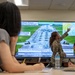 A military briefing is taking place in a conference room where a U.S. Air Force service member in camouflage uniform is presenting a slide to a mixed audience of military personnel and civilians. The presentation screen displays a slide titled 'The Shogun Mission: Deterrence Through Strength' with bilingual English and Japanese text, showing an airfield with military aircraft and a regional map. The presenter is pointing to a map in the upper right corner of the screen that appears to show East Asia with range circles.