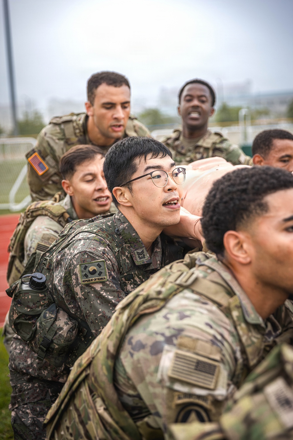 A group of military personnel from multiple nations are engaged in a joint training exercise, appearing to carry or lift a heavy object or person together. At least one soldier is visibly wearing a 'Republic of Korea' patch with the South Korean flag emblem, while others wear US Army MultiCam/OCP uniforms with American flag patches. The setting appears to be an outdoor athletic track facility.