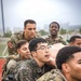A group of military personnel from multiple nations are engaged in a joint training exercise, appearing to carry or lift a heavy object or person together. At least one soldier is visibly wearing a 'Republic of Korea' patch with the South Korean flag emblem, while others wear US Army MultiCam/OCP uniforms with American flag patches. The setting appears to be an outdoor athletic track facility.