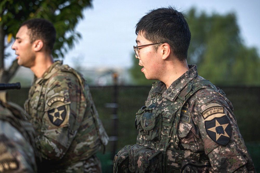 Two soldiers in military uniforms are visible, one wearing US Army OCP (Operational Camouflage Pattern) and one wearing Republic of Korea Army (ROKA) digital camouflage. Both display the 2nd Infantry Division patch on their left shoulders, indicating this is a Combined Division joint unit. The foreground soldier wearing glasses has a 'ROKA' and 'Combined Division' patch visible, suggesting a US-South Korea joint military exercise or operation.