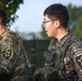 Two soldiers in military uniforms are visible, one wearing US Army OCP (Operational Camouflage Pattern) and one wearing Republic of Korea Army (ROKA) digital camouflage. Both display the 2nd Infantry Division patch on their left shoulders, indicating this is a Combined Division joint unit. The foreground soldier wearing glasses has a 'ROKA' and 'Combined Division' patch visible, suggesting a US-South Korea joint military exercise or operation.